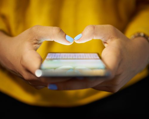 Young woman using cell phone to send text message on social network at night. Closeup of hands with computer laptop in background